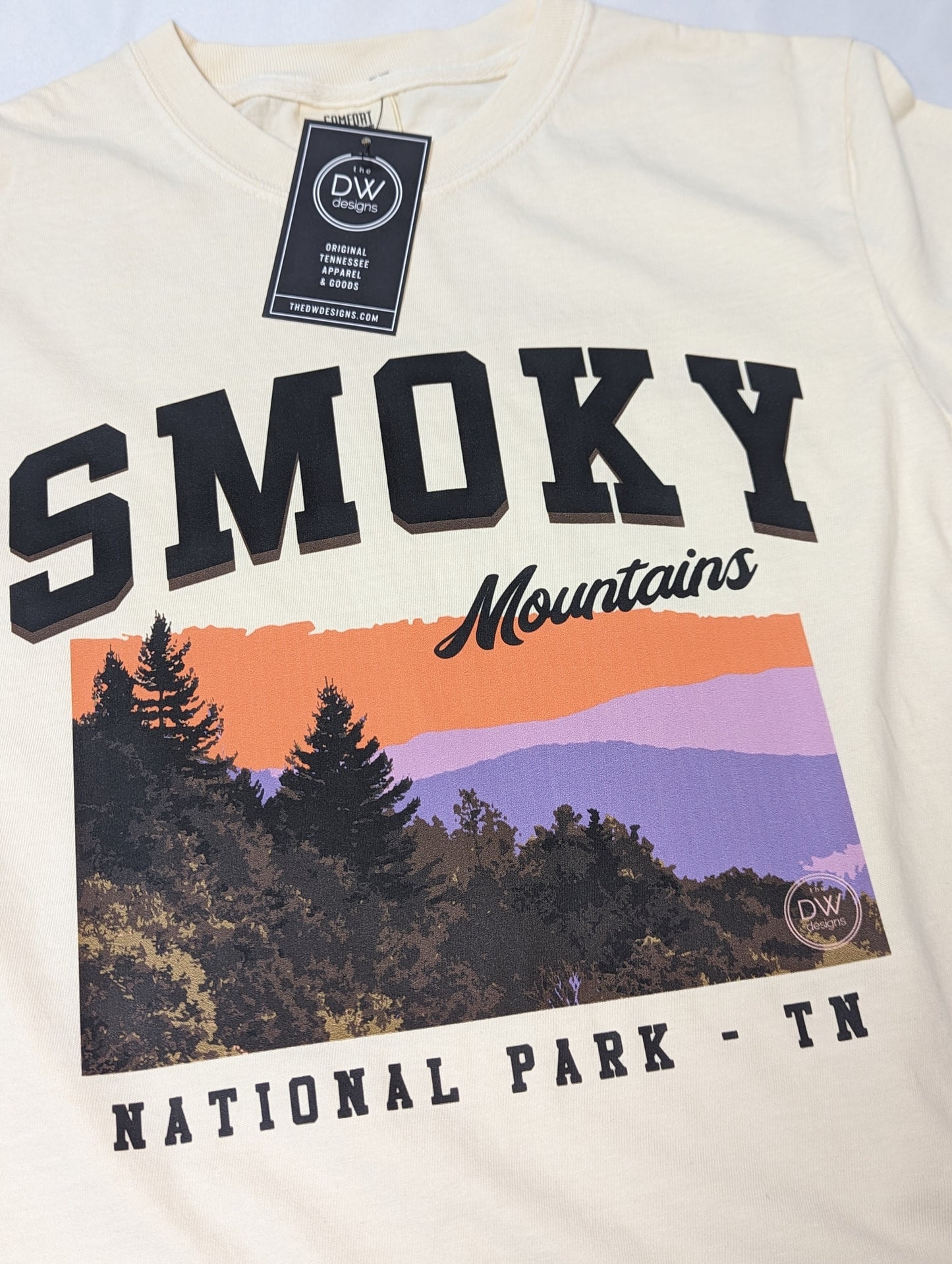 The Smoky Mountains Graphic Tee features a colorful mountain sunset above the trees in the Smoky Mountains National Park, printed on an off-white t-shirt. Sold by The DW Designs, Knoxville, TN.